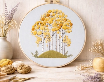 May include: Hand-stitched embroidery art in a wooden hoop. The design features a cluster of birch trees with yellow leaves on a green hill. The trees have white bark with black markings. The embroidery is set against a white background.