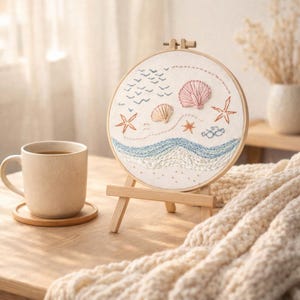 May include: A round embroidery hoop featuring a beach scene with seashells, starfish, and birds. The artwork is displayed on a small wooden easel. A beige mug of tea sits on a wooden coaster, with a cream-colored blanket in the foreground.