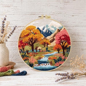May include: A circular embroidery hoop displaying a landscape scene with a river, trees, and mountains. The trees are in autumnal shades of orange, red, and yellow. The mountains are blue and white, and the river is blue.