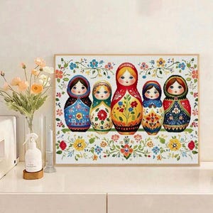 May include: A framed cross-stitch depicting five traditional Matryoshka dolls. The dolls are brightly coloured with floral patterns and are set against a white background with a floral border. The dolls are arranged in a row.