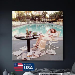May include: A canvas print featuring a vintage photograph of a person relaxing by a pool, surrounded by newspapers and an Oscar statue. The image is set in a luxurious outdoor setting, with a pool and palm trees in the background. The print includes the text "Made in USA" and the Sea Wall Arts logo.