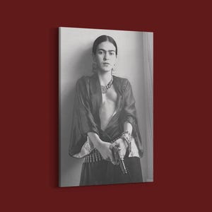 May include: Black and white portrait print of a woman in a dark robe, holding a gun. She wears a necklace, earrings, and bracelets. The image is a canvas print, leaning against a wall.