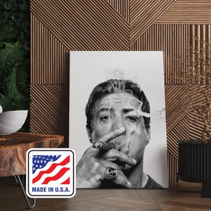 Puede incluir: Impresión en lienzo en blanco y negro que presenta un retrato de un hombre fumando un cigarro. La imagen muestra el rostro del hombre y su mano sosteniendo el cigarro, con humo ascendente. La impresión tiene una pegatina "MADE IN U.S.A.".