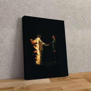 Puede incluir: Impresi&oacute;n en lienzo enmarcada que muestra a dos figuras d&aacute;ndose la mano sobre un fondo negro. Una figura est&aacute; en llamas, la otra con un traje verde oscuro. La obra de arte se exhibe sobre una superficie de madera.