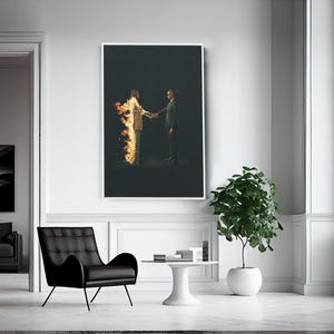 Puede incluir: Una impresi&oacute;n art&iacute;stica enmarcada muestra a dos figuras d&aacute;ndose la mano sobre un fondo oscuro. Una figura est&aacute; en llamas, la otra con traje. La obra de arte est&aacute; en una habitaci&oacute;n blanca con una silla negra y una planta en maceta.