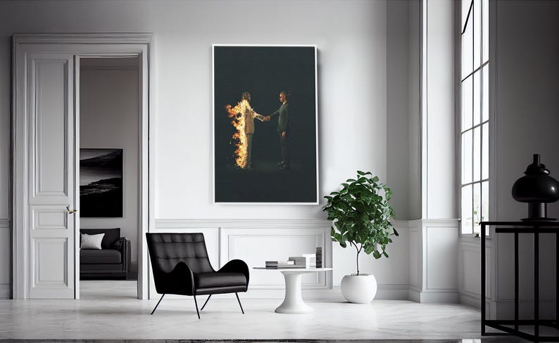 Puede incluir: Una impresi&oacute;n art&iacute;stica enmarcada muestra a dos figuras d&aacute;ndose la mano sobre un fondo oscuro. Una figura est&aacute; en llamas, la otra con traje. La obra de arte est&aacute; en una habitaci&oacute;n blanca con una silla negra y una planta en maceta.