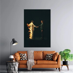 Puede incluir: Una gran impresi&oacute;n art&iacute;stica enmarcada representa a dos figuras d&aacute;ndose la mano. Una figura est&aacute; en llamas, la otra con un traje. La obra de arte se exhibe sobre un sof&aacute; de cuero marr&oacute;n con cojines y una manta. Tambi&eacute;n se ve una l&aacute;mpara de pie negra y una mesa auxiliar.