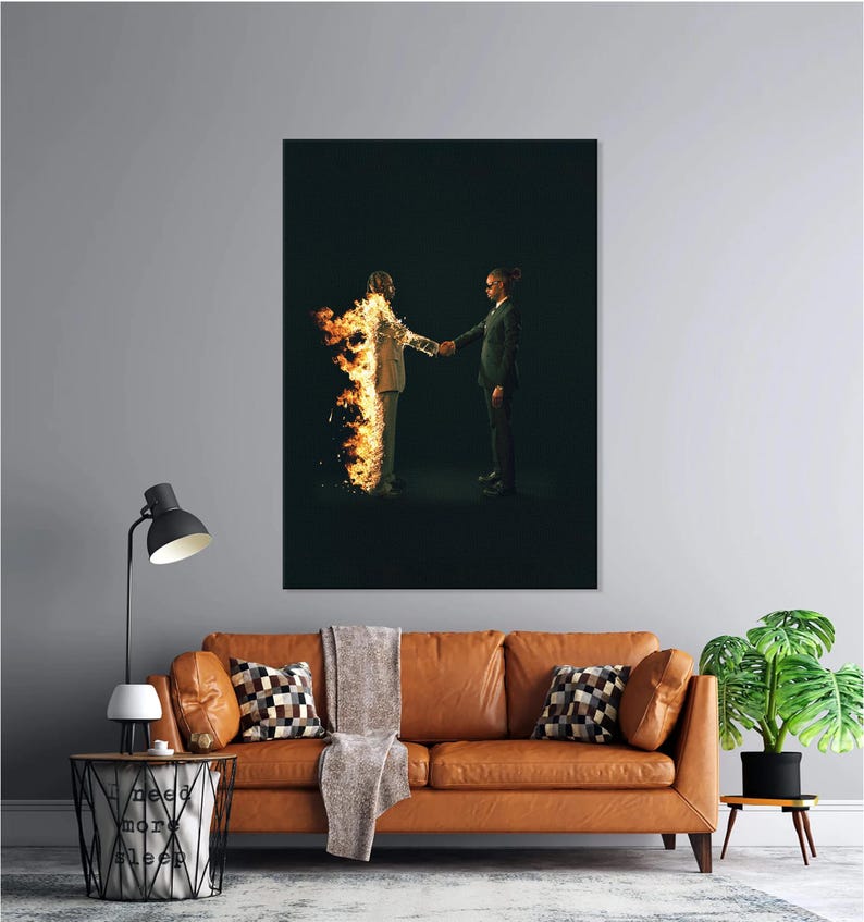 Puede incluir: Una gran impresi&oacute;n art&iacute;stica enmarcada representa a dos figuras d&aacute;ndose la mano. Una figura est&aacute; en llamas, la otra con un traje. La obra de arte se exhibe sobre un sof&aacute; de cuero marr&oacute;n con cojines y una manta. Tambi&eacute;n se ve una l&aacute;mpara de pie negra y una mesa auxiliar.