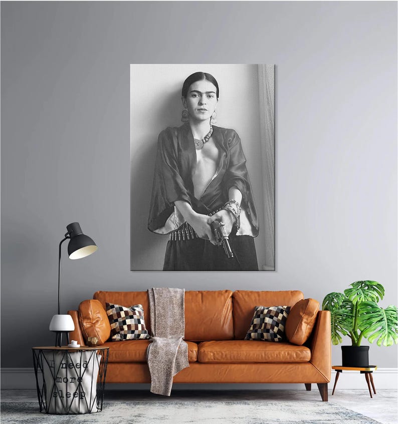 May include: Black and white portrait of a person holding a gun, wearing jewelry and a dark top. The image is a print, likely a photograph, and is displayed on a wall above a brown leather sofa. The room has a modern aesthetic.