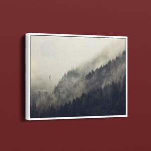 May include: Framed canvas art print featuring a misty mountain scene. The artwork depicts a forest on a hillside, shrouded in fog, with a soft, pale sun in the sky. A bird is in flight. The artwork is framed in a white border.