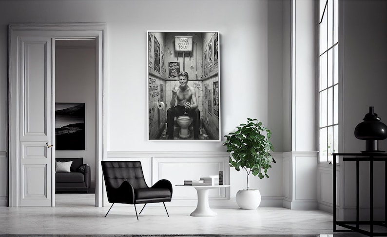 Peut inclure: Impression en noir et blanc d'un homme assis sur des toilettes, entour&eacute; d'affiches. La pi&egrave;ce est lumineuse, avec une chaise noire et une petite table. Une plante en pot et une fen&ecirc;tre sont visibles.