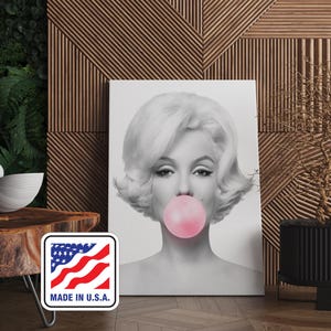 Könnte beinhalten: Schwarz-Weiß-Porträt von Marilyn Monroe mit einer rosa Kaugummiblase. Das Kunstwerk befindet sich auf einer weißen Leinwand vor einem Holz Hintergrund. Ein "Made in U.S.A." Aufkleber ist unten links zu sehen.