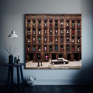 May include: A framed print of a brownstone building with numerous windows, each occupied by a person in a colorful dress, arms raised. A vintage car is parked in front of the building. The artwork is displayed on a light blue wall.