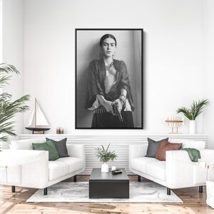 May include: Black and white framed print of Frida Kahlo. The portrait shows Kahlo in a dark top and skirt, with jewelry and a serious expression. The image is displayed above a white couch.