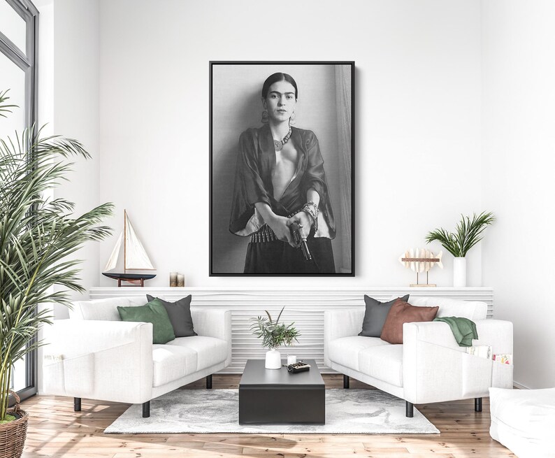 May include: Black and white framed print of Frida Kahlo. The portrait shows Kahlo in a dark top and skirt, with jewelry and a serious expression. The image is displayed above a white couch.