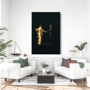 Puede incluir: Una impresi&oacute;n art&iacute;stica enmarcada representa a dos figuras d&aacute;ndose la mano sobre un fondo negro. Una de las figuras est&aacute; envuelta en llamas. La obra de arte se exhibe sobre un sof&aacute; blanco con cojines verdes y grises, una mesa de centro negra y una planta en maceta.