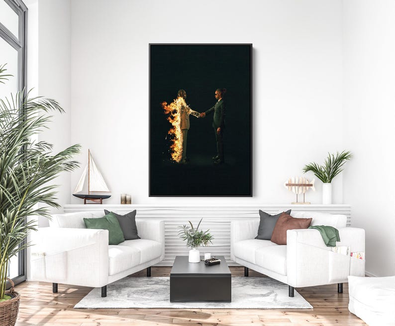 Puede incluir: Una impresi&oacute;n art&iacute;stica enmarcada representa a dos figuras d&aacute;ndose la mano sobre un fondo negro. Una de las figuras est&aacute; envuelta en llamas. La obra de arte se exhibe sobre un sof&aacute; blanco con cojines verdes y grises, una mesa de centro negra y una planta en maceta.