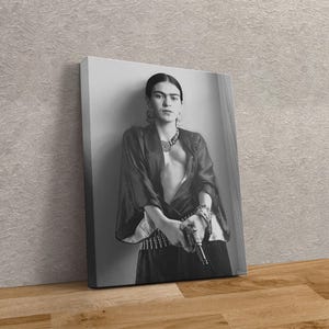 May include: Black and white canvas print featuring a portrait of Frida Kahlo holding a gun. She is wearing a dark top, jewelry, and a skirt. The print is leaning against a wall.