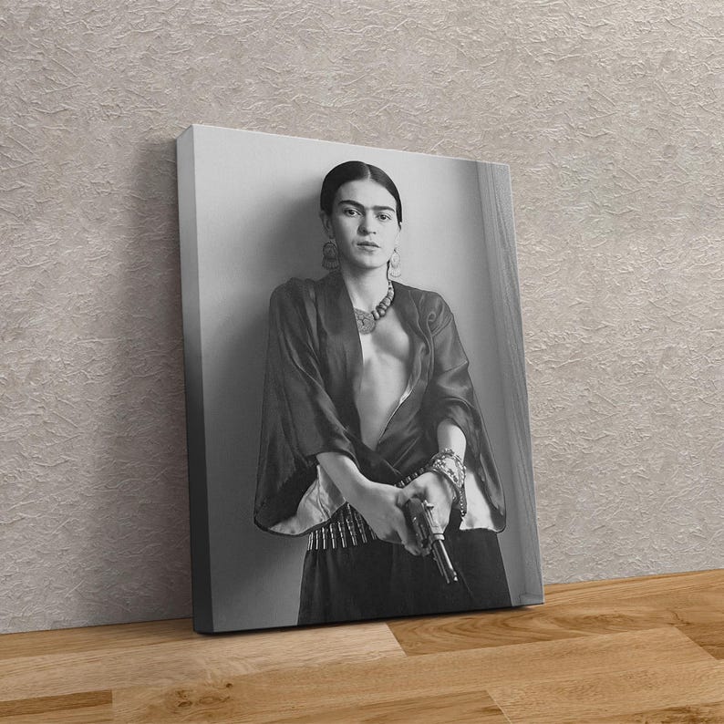 May include: Black and white canvas print featuring a portrait of Frida Kahlo holding a gun. She is wearing a dark top, jewelry, and a skirt. The print is leaning against a wall.
