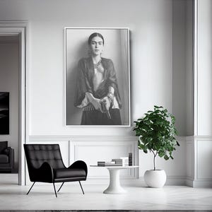 May include: A black and white framed portrait of a woman hangs on a white wall. A black armchair, a small white table, and a potted plant are in front of the wall. A window with a black frame is on the right.