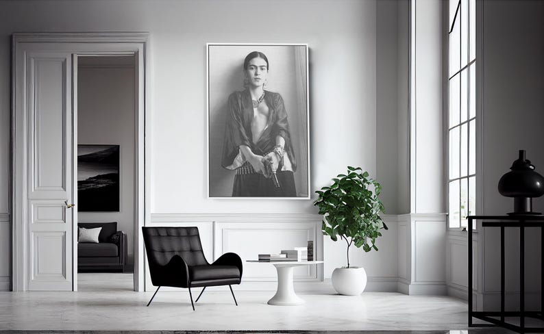 May include: A black and white framed portrait of a woman hangs on a white wall. A black armchair, a small white table, and a potted plant are in front of the wall. A window with a black frame is on the right.