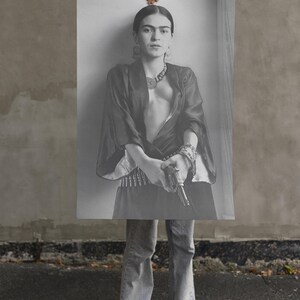 May include: Black and white print of a woman holding a gun, wearing a dark top, jewelry, and jeans. The image is held up by a hand, with the woman's lower body and feet visible. The background is a gray wall and asphalt.