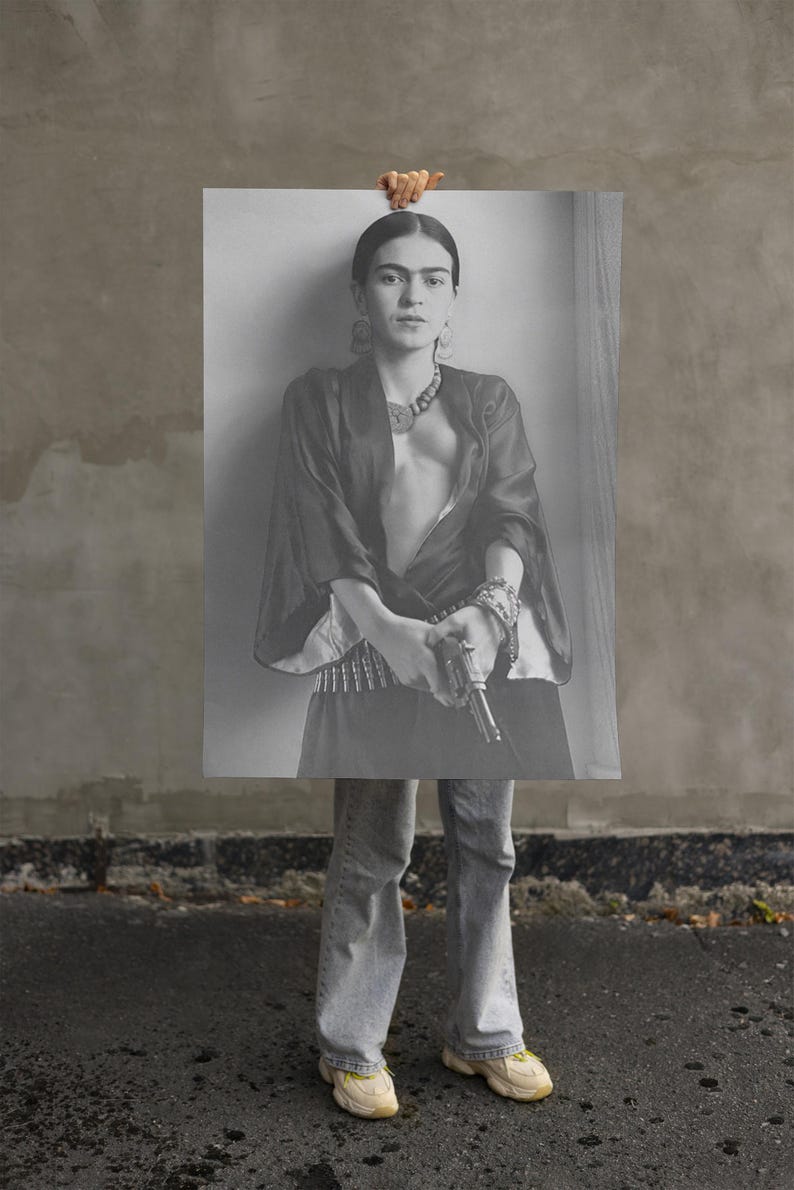 May include: Black and white print of a woman holding a gun, wearing a dark top, jewelry, and jeans. The image is held up by a hand, with the woman's lower body and feet visible. The background is a gray wall and asphalt.