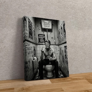 Peut inclure: Impression sur toile en noir et blanc montrant une personne assise sur des toilettes, levant un doigt et tenant un verre. Les murs sont couverts d'affiches et de graffitis, avec les textes "SPACE ODDITY TOILET" et "ZIGGY STARDUST WAS HERE".