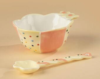 May include: A ceramic baby bowl and spoon set. The bowl is yellow with pink and polka dot accents. The spoon is yellow with a flower-shaped bowl and a wavy handle. The set is designed for serving baby food.