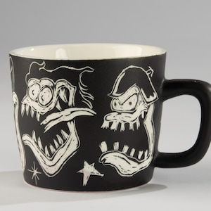 Può includere: Tazza in ceramica nera con interno bianco e manico nero. La tazza presenta illustrazioni bianche di facce di mostri dei cartoni animati. Una stella bianca è visibile anche all'esterno della tazza.