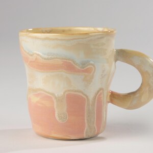 Pode incluir: Uma caneca de cerâmica artesanal com uma forma única e orgânica. A caneca apresenta uma mistura de tons pêssego, creme e azul claro, com efeitos de esmalte escorrido. A alça é um laço arredondado, adequada para bebidas quentes ou frias.
