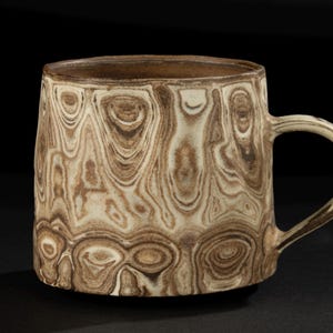 Puede incluir: Una taza de cerámica con un patrón único de vetas de madera en tonos marrón y crema. La taza tiene un cuerpo redondeado, un asa robusta y un borde marrón oscuro. El diseño le da a la taza una estética natural y terrosa.