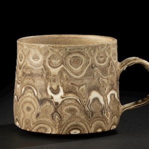 Può includere: Una tazza in ceramica con un motivo marmorizzato unico nei toni del marrone e del crema. La tazza ha un corpo arrotondato, un manico robusto ed è adatta per caffè o tè. Il design conferisce alla tazza un aspetto rustico e artigianale.