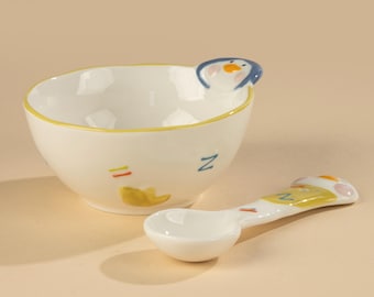 May include: A white ceramic bowl and spoon set. The bowl has a yellow rim and a penguin design on the side. The spoon has a matching penguin design. The bowl has a yellow duck design and the letter "Z".