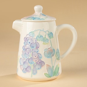 Könnte beinhalten: Eine weiße Keramik-Teekanne mit Deckel, Henkel und Ausguss. Die Teekanne ist mit einem Blumendesign in Blau-, Lila- und Grüntönen verziert. Die Blumen umfassen Traubenhyazinthen und Tulpen.