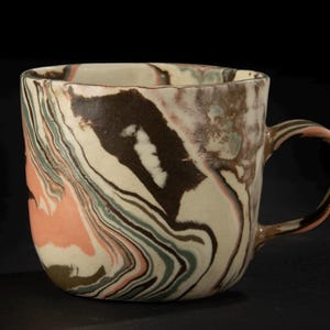 Peut inclure: Une tasse en céramique avec un motif marbré unique. La tasse présente un mélange de couleurs, notamment du marron, crème, rose et vert, créant un motif tourbillonnant. Elle a un corps arrondi, une anse et un bord carré.