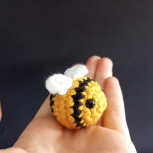Peut inclure: Une petite abeille au crochet faite à la main, avec des rayures jaunes et noires. L'abeille a des ailes blanches et des yeux en perles noires. L'abeille est tenue dans une main sur un fond sombre. Il s'agit d'un artisanat mignon et fait à la main.