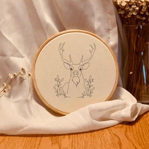 Pode incluir: Um bastidor de bordado redondo com um desenho a traço preto de um veado com chifres e detalhes florais. A obra de arte está em tecido creme, num bastidor de madeira natural. Flores secas num vaso de vidro ao fundo.
