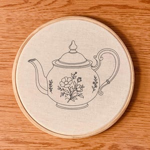 Pode incluir: Design de bule bordado em tecido creme, esticado num bastidor de bordado de madeira. O bule apresenta padrões florais e de folhas, feitos com fio preto. O fundo é uma superfície de madeira.