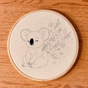 Pode incluir: Um bastidor de bordado redondo com um coala bordado à mão e um desenho floral. O coala está sorrindo, e as flores e folhas estão em um delicado estilo de desenho a traço. O tecido é de cor bege claro, e o bastidor é de madeira.