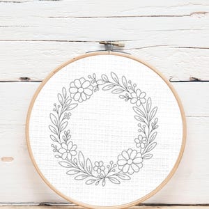 Pode incluir: Um bastidor de bordado floral com um desenho de grinalda. A grinalda é composta por flores e folhas estilizadas, costuradas com fio cinzento sobre tecido branco. O bastidor de madeira é redondo e tem um fecho de metal.
