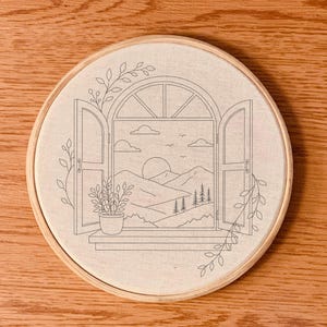 May include: A circular wooden embroidery hoop containing a piece of off-white fabric with a black line art design of an open window looking out onto a landscape with mountains, a sun, clouds, trees, and a potted plant on the windowsill.