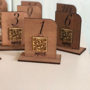 Può includere: Segnaposto in legno con la parte superiore curva e una base rettangolare. Ogni segnaposto ha un numero, un codice QR e la parola "Menu". I segnaposto sono in legno marrone chiaro e sono disposti su una superficie bianca.