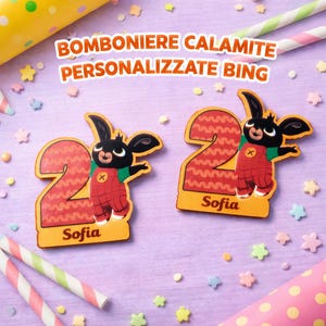 Può includere: Due calamite personalizzate a tema Bing con il numero 2, raffiguranti un personaggio di coniglio dei cartoni animati con una tuta rossa. Le calamite sono su una superficie viola con coriandoli colorati e cannucce a righe. Il nome "Sofia" è stampato.