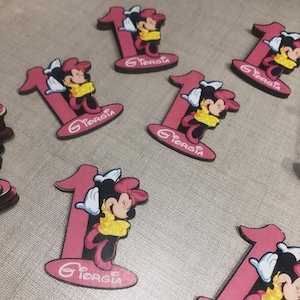 Puede incluir: Recortes rosas con el número uno y gráficos de Minnie Mouse. Cada recorte presenta un diseño de Minnie Mouse con el nombre "Giorgia" en cursiva. Los recortes son probablemente para una celebración de primer cumpleaños.