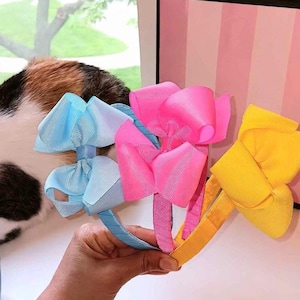 May include: Three hair headbands featuring large, multi-layered grosgrain ribbon bows in solid light blue, bright pink, and yellow. Each headband is wrapped in matching ribbon, showcasing a textured, ribbed fabric finish on both the bows and bands.