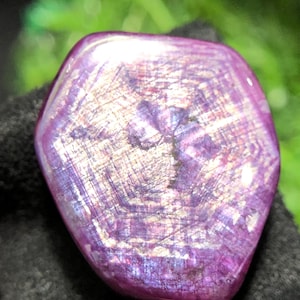 Puede incluir: Una gema hexagonal pulida con un vibrante tono púrpura. La superficie de la piedra muestra un patrón brillante e iridiscente con toques de plata y oro. La gema se encuentra sobre un fondo oscuro, resaltando su color y textura.