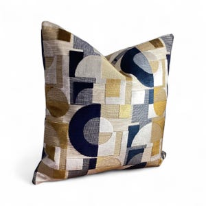 May include: Decorative throw pillow with a geometric pattern. The pillow features a repeating design of navy blue, gold, beige, and white shapes. The shapes include circles, squares, and rectangles. The pillow has a textured appearance.