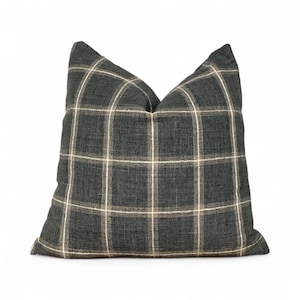 May include: A square decorative pillow with a plaid pattern. The pillow features a dark gray fabric with a grid of white and tan lines. The pillow is a neutral color and would complement a variety of home decor styles.