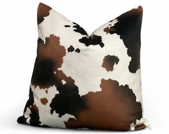 May include: A square decorative pillow with a cowhide pattern. The pillow features a mix of white, black, and brown patches, creating a rustic and natural aesthetic. The pillow is designed for home decor.